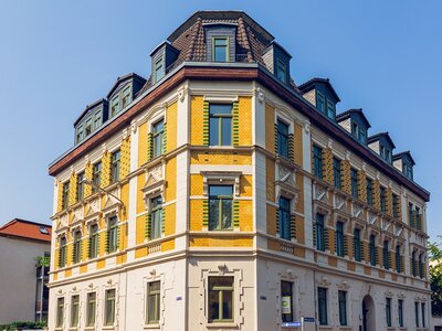 Historisches Eckgebäude mit gelber und weißer Fassade sowie grünen Fensterläden unter klarem, blauem Himmel