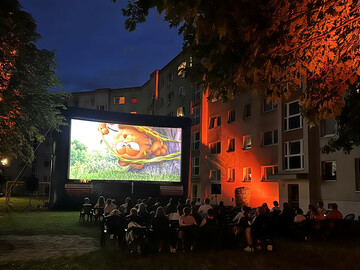 Menschen sitzen beim Sommerkino zusammen. Auf einer großen Leinwand läuft Garfield der Film.