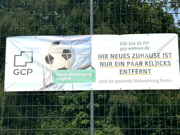 An einem Zaun hängt ein Banner von GCP mit der Aufschrift "Ihr neues Zuhause ist nur ein paar Klicks entfernt."