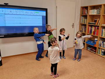 Fünf Kinder stehen vor einem großen Bildschirm, wo Tonspuren zu sehen ist. Zwei Kinder haben Mikrofone in der Hand und alle Kinder singen. 