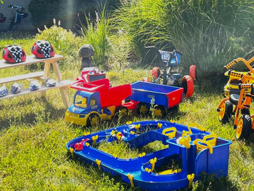 Auf einer Wiese steht verschiedenes Spielzeug. Darunter Kinderfahrräder, ein Spielzeug-Laster und eine Kinderwasserbahn. Auf einer Bank sind Fahrradhelme für Kinder aufgereiht. 
