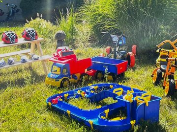 Auf einer Wiese steht verschiedenes Spielzeug. Darunter Kinderfahrräder, ein Spielzeug-Laster und eine Kinderwasserbahn. Auf einer Bank sind Fahrradhelme für Kinder aufgereiht. 