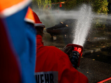 Ein Junge als Feuerwehrmann, der von Hinten zu sehen ist, spritzt mit einem Wasserstrahl in Richtung eines Feuers. 